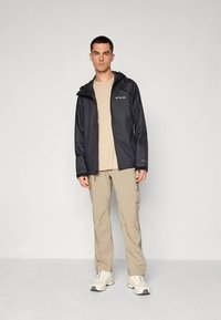 Musta vedenpitävä takki hupulla, vetoketjulla edessä ja logolla rinnassa, yhdistetty beigeihin housuihin ja valkoisiin lenkkareihin. Sileä pinta.