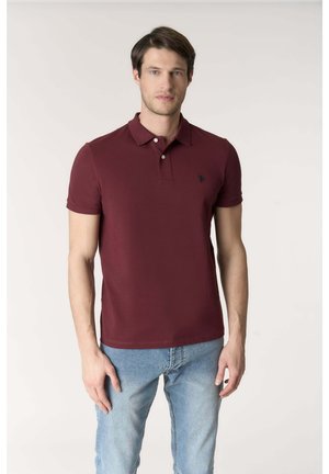 Giovane uomo in piedi che indossa una polo bordeaux a maniche corte e jeans azzurri davanti a uno sfondo chiaro e uniforme.