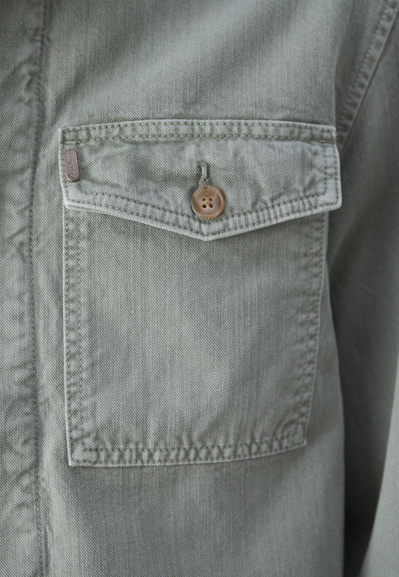 Camisa de mezclilla verde con un bolsillo en el pecho que cuenta con un cierre de botón marrón y una pequeña etiqueta de metal. Tejido texturizado con un diseño casual.