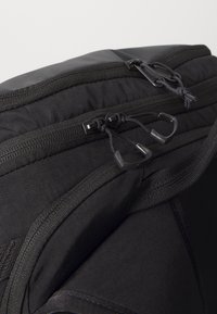 Gros plan de la partie supérieure d’un sac à dos noir montrant trois tirettes de fermeture éclair avec des boucles de corde sur une surface en tissu résistante.