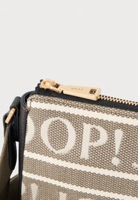 Béžová kanvasová crossbody taška s bílým nápisem. Má zlaté kování, horní zipové zapínání a texturovaný design tkaniny.