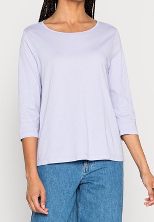 Kobieta ubrana w jasnofioletową bluzkę z rękawami do łokcia i niebieskie dżinsy, stojąca na jednolitym tle.