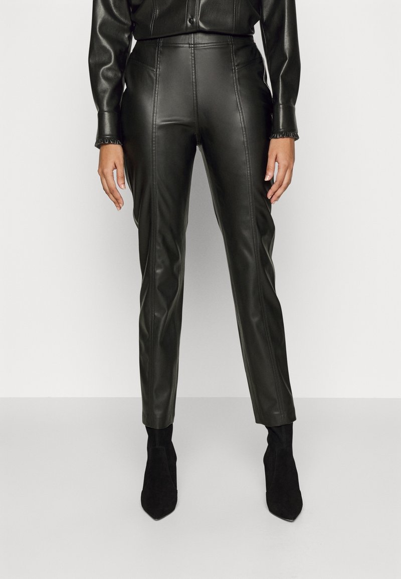 Femme portant un pantalon noir en faux cuir taille haute associé à des bottines noires à bout pointu sur un fond clair uni.
