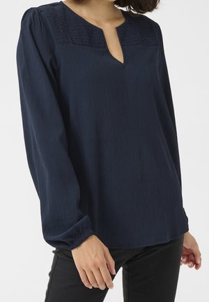Femme portant un chemisier bleu foncé à manches longues avec un panneau supérieur texturé et une petite ouverture en V, assorti à un pantalon noir.