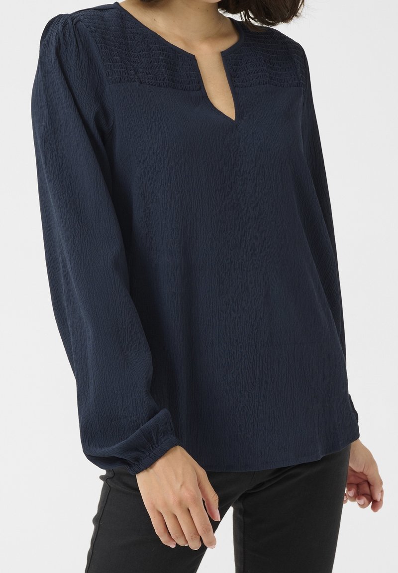 Femme portant un chemisier bleu foncé à manches longues avec un panneau supérieur texturé et une petite ouverture en V, assorti à un pantalon noir.