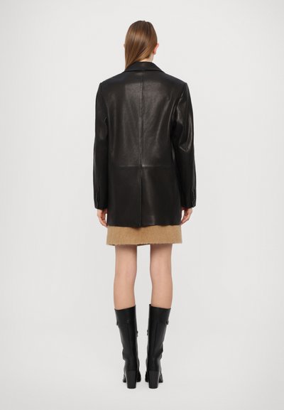 Sieviete stāv ar muguru pret skatītāju, ģērbta melnā ādas žaketē, gaišā svārku garumā un melnās ceļgalu augstās zābakos, fona ir vienkāršs.