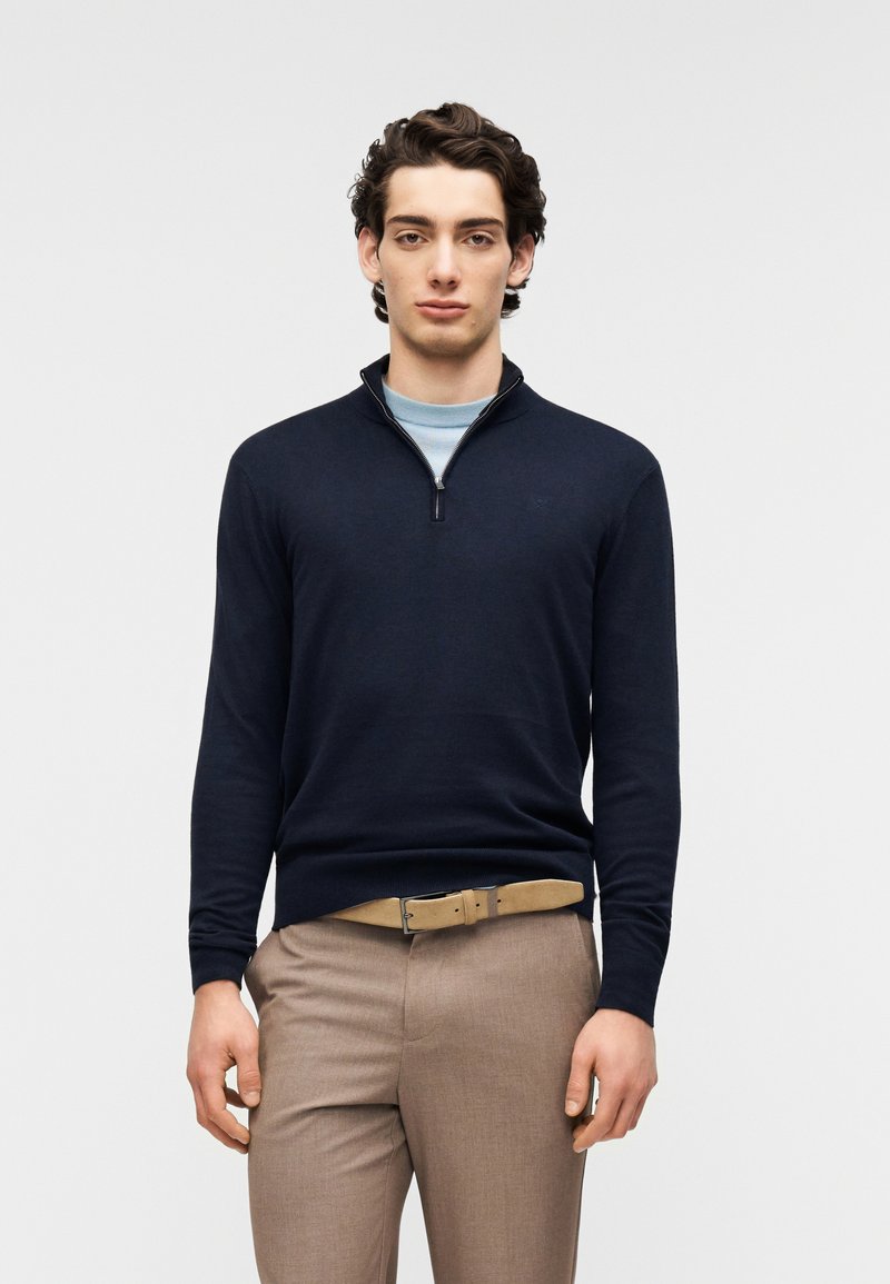 Pull à col zippé bleu marine à manches longues, en tissu tricoté, porté sur un sous-pull bleu clair, assorti à un pantalon beige et une ceinture.
