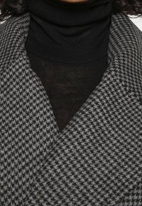 Grijze houndstooth jas van een textuurrijke, zachte stof, met een grote kraag en gedragen over een zwart coltrui.