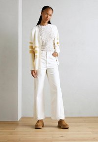 Cardigan crema con strisce gialle sopra una maglia bianca testurizzata, abbinato a pantaloni bianchi a gamba larga e scarpe chunky marroni, in piedi su un pavimento di legno.