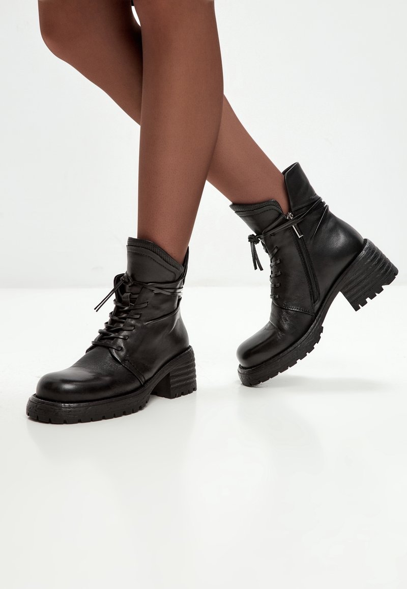 Jambes à la peau foncée portant des bottines en cuir noir à lacets avec fermetures éclair latérales et talons épais, debout sur un sol blanc.