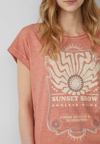 Pale coral t-shirt met kapmouwtjes. Centraal grafisch ontwerp bevat een zon en de tekst "SUNSET SHOW" in goud met een versleten textuur.