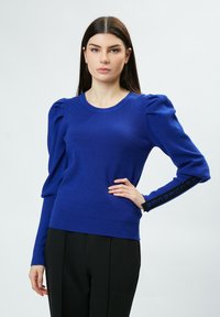 Blauer Strickpullover mit Puffärmeln und rundem Halsausschnitt, mit seitlichen Schlitzen und schwarzen Textakzenten an den Manschetten. Glatte Textur.