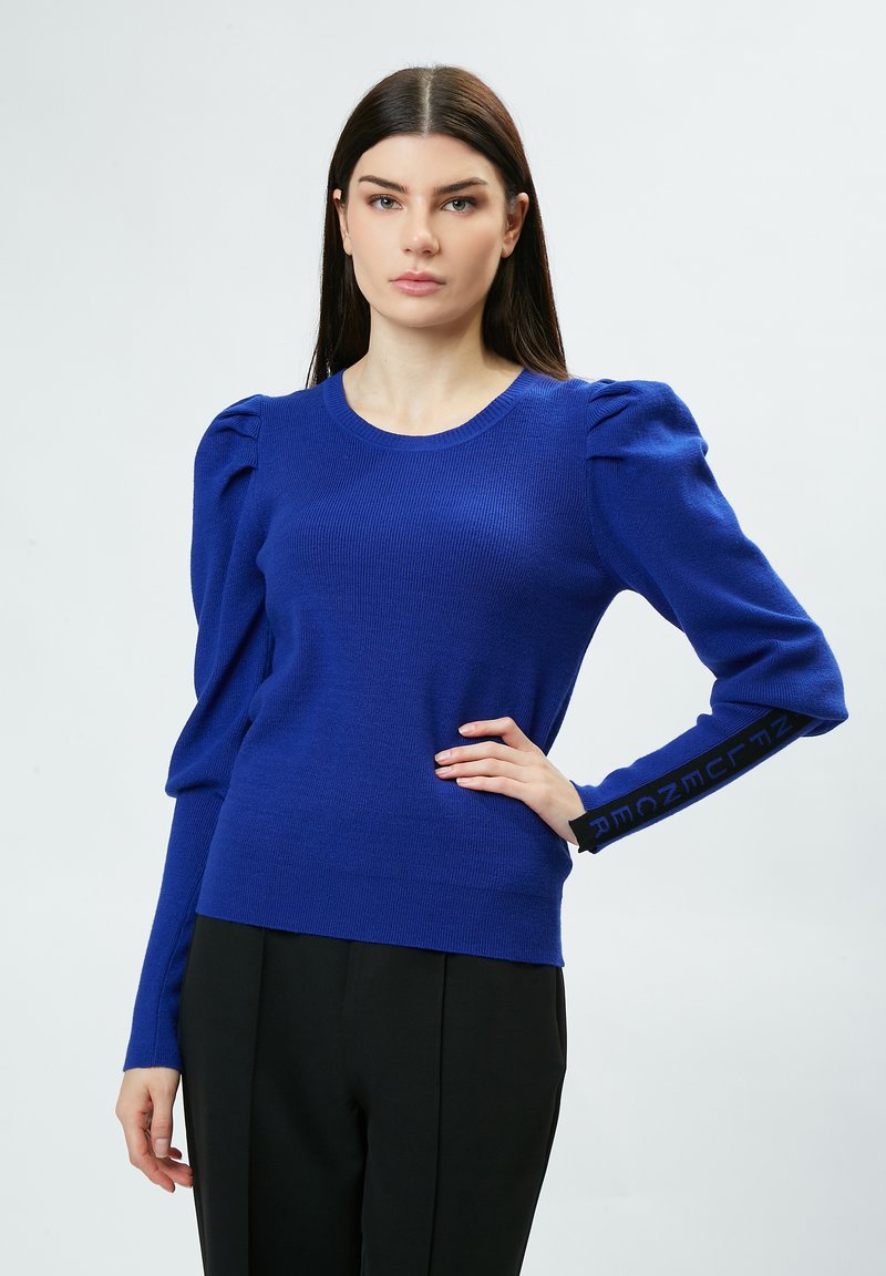 Blauer Strickpullover mit Puffärmeln und rundem Halsausschnitt, mit seitlichen Schlitzen und schwarzen Textakzenten an den Manschetten. Glatte Textur.