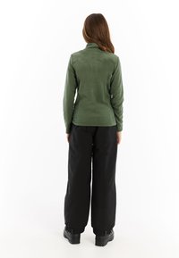 Grüne Fleecejacke mit hohem Kragen, kombiniert mit schwarzen weit geschnittenen Hosen und robusten schwarzen Stiefeln, von hinten auf einem weißen Hintergrund betrachtet.