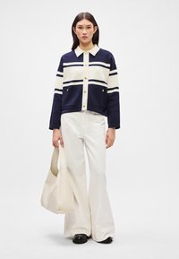 Jūrinės (navy) ir kreminės (cream) juostelės megztinis su apykakle, auksiniais mygtukais ir priekinėmis kišenėmis, derinamas su kreminėmis (cream) plačiomis kelnių ir derančia rankine.