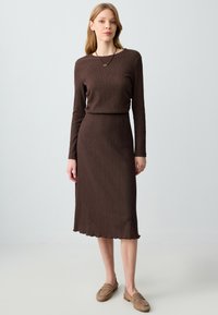 Robe longue à manches longues de couleur brune avec une taille cintrée, un ourlet évasé et un motif côtelé. Associée à des chaussures à enfiler de couleur brun clair.