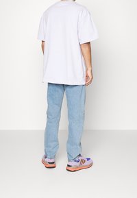 T-shirt ample lavande clair, jean en denim bleu clair, et baskets multicolores avec des accents violets, gris et orange, vus de dos.