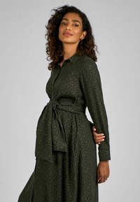 Abito verde a maniche lunghe con stampa leopardata, vita con cintura, colletto e tessuto fluente. Presenta dettagli con bottoni e una vestibilità ampia e comoda.