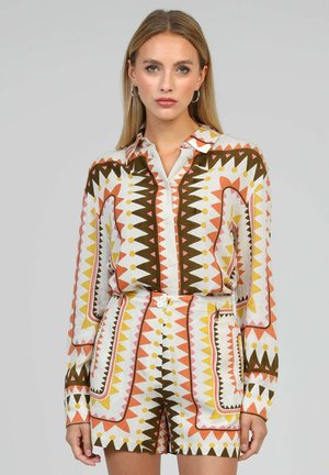 Femme portant une chemise à manches longues et un short assorti avec un motif géométrique en zigzag orange, jaune, marron et blanc sur fond uni.