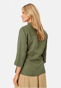 Olivgrünes Hemd mit Dreiviertelärmeln, lässiger Passform und einer einzigen Hinterkante Naht. Glatte Textur, mit einem Saum, der hinten leicht länger ist.