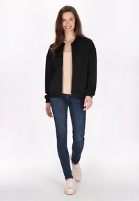 Zwarte bomberjack met een visgraatpatroon, ritssluiting, geribde manchetten en zoom, gecombineerd met een beige top en blauwe jeans. Lichte sneakers.