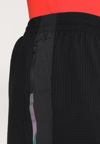 Pantalones cortos deportivos negros con una superficie texturizada, que cuentan con un bolsillo con cremallera y una franja multicolor a lo largo del costado. Cintura elástica.