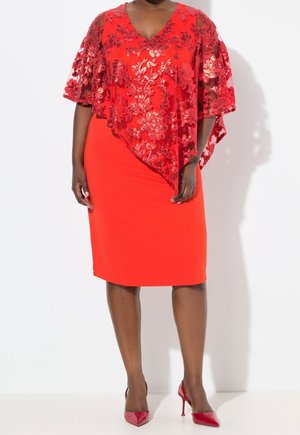 Kvinde iført en knallød knælang kjole med gennemsigtigt blomstermønster i blonder og matchende røde højhælede sko foran en hvid baggrund.