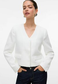 Bílý zkrácený blazer s texturovaným povrchem, výstřihem do V a dvěma předními kapsami, zkombinovaný s tmavými džíny. Dlouhé rukávy a precizní střih.