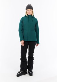 Mujer vestida con chaqueta de esquí verde oscuro, pantalones de esquí negros, botas de esquí negras y un gorro gris, de pie contra un fondo blanco.