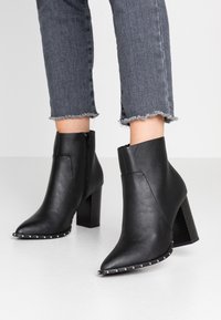 Stivaletti neri in pelle con punta appuntita, tacco a blocco di 7 cm, zip laterale e dettagli borchiati sulla suola. Indossati con jeans con orlo sfrangiato.