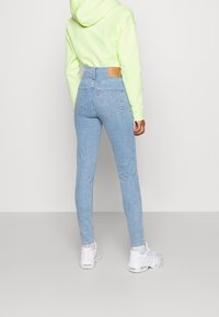 Jeans de tiro alto en azul claro con un parche de cuero marrón en la parte posterior y un diseño de bolsillo minimalista, combinados con una sudadera verde brillante.