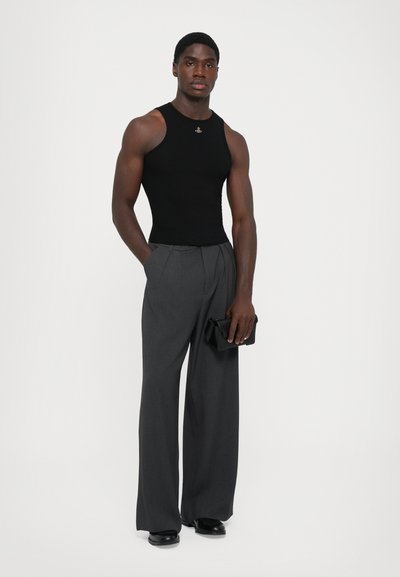 Man in een zwart mouwloos shirt, donkergrijze wijde broek, zwarte dresschoenen, met een kleine zwarte clutch in de hand, staand tegen een witte achtergrond.