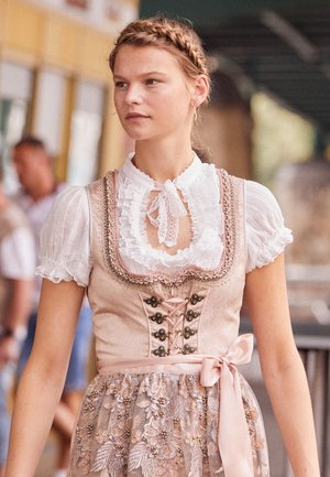 Junge Frau mit geflochtenem Haar trägt ein beiges Dirndl mit Spitzenbluse und floral besticktem Schürzen, steht im Freien.
