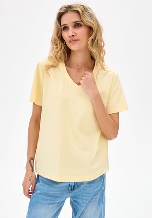 Blondynka o falujących włosach, ubrana w bladożółtą koszulkę z dekoltem w serek i jasnoniebieskie dżinsy, stojąca na tle białego tła.