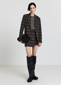 Brauner kariert Blazer über einem grauen strukturierten Pullover, kombiniert mit einem karierten Minirock und kniehohen schwarzen Lederstiefeln. Hält eine schwarze Clutch.