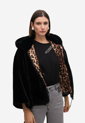 Giacca in pelliccia finta nera con rivestimento in stampa leopardata, cappuccio oversize, chiusura con zip e top nero abbinato a jeans grigi.