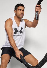 Fehér ujjatlan felső fekete Under Armour logóval, fekete sort és egy textúrált fekete harci kötelet tartanak fókuszált fogásban. Semleges háttér.