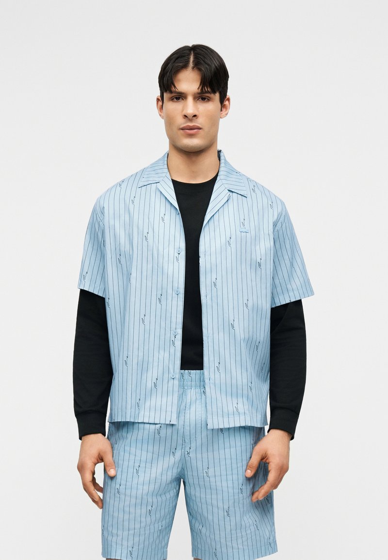 Homme portant une chemise à manches courtes rayée bleu clair et un short assorti par-dessus un haut noir à manches longues, debout devant un fond uni.