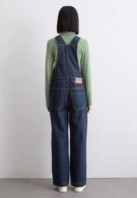 Džínové šortky s širokými nohavicemi, s nastavovacími popruhy, hnědou nášivkou na zadní straně a dvěma zadními kapsami, nošené přes top s zelenými proužky.