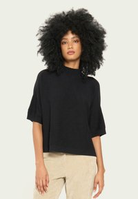 Pulover din tricot negru, cu un guler înalt și mâneci scurte largi, asortat cu pantaloni din corduroy de culoare deschisă.