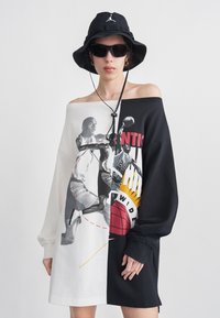 Άτομο που φοράει υπερμεγέθη μαύρο και λευκό φόρεμα εκτός ώμου με εκτύπωση παίκτη μπάσκετ, μαύρο καπέλο τύπου bucket και σκοτεινά γυαλιά ηλίου.