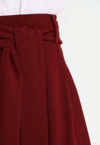 Jupe bourgogne avec une large ceinture à nouer et un design plissé. Fabriquée en tissu doux avec une texture lisse et sans motifs visibles.