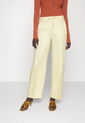 Vrouw draagt lichtgele wijdvallende broek met aantrekkoord in de taille, roestkleurige geribbelde top met lange mouwen en een sleutelgat, en mosterdkleurige sandalen met hoge hakken.
