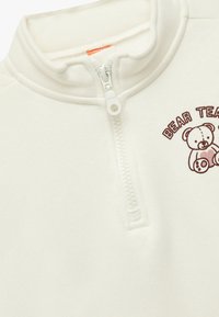 Valkoinen fleece-puolipitkä takki, jossa on mock neck -kaulus. Siinä on ruskea karhun grafiikka ja "BEAR TEA" -teksti. Pehmeä pinta ja hienovaraiset ompeluyksityiskohdat.