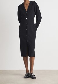 Robe noire en maille à manches longues avec un décolleté en V, boutonnage sur le devant et fente en bas. Associée à des chaussures Oxford noires avec des détails en dentelle.