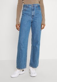 Person klädd i högmidjade vida blå jeans, beige långärmad topp och vita träningsskor, stående mot en enkel bakgrund.