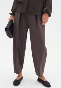 Brown wide-leg trousers made of smooth fabric, featuring a high waist and subtle pleats, paired with black flats and a black clutch.