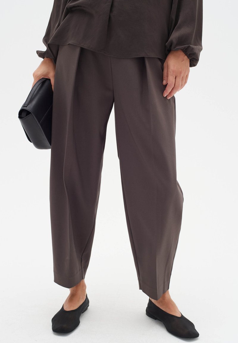 Brown wide-leg trousers made of smooth fabric, featuring a high waist and subtle pleats, paired with black flats and a black clutch.