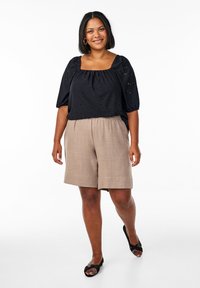 Top nero con occhietti e maniche a sbuffo, scollatura quadrata, abbinato a pantaloni corti di lino beige. Il modello indossa sandali neri con accenti decorati.