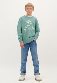 Felpa verde menta con un design grafico di un teschio; abbinata a jeans azzurri arrotolati e sneaker bianche con accenti beige.